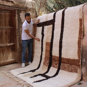 May include: A large, rectangular rug with a cream-colored background and a geometric pattern in shades of brown and black. The rug has a thick, textured pile and a fringe along one edge. The rug is displayed outdoors.