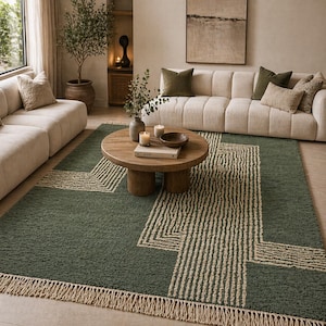 Pu&ograve; includere: Un tappeto verde con un motivo geometrico beige e bordi con frange. Un tavolino rotondo in legno &egrave; posto sul tappeto. Il soggiorno presenta un divano color crema e cuscini.