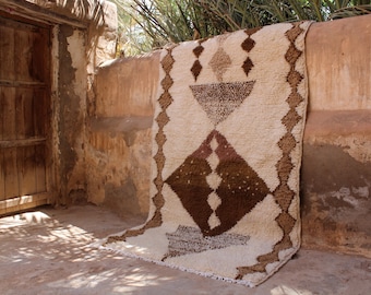 May include: A beige and brown geometric patterned rug is draped over a weathered wall. The rug features diamond and triangle shapes in shades of brown against a beige background. The rug's fringe hangs down, and the scene is set in a rustic outdoor space.