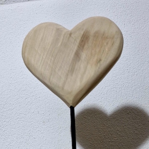 Könnte beinhalten: Eine Holzherzskulptur auf einem schwarzen Metallständer. Das Herz ist hellfarbig und der Ständer hat einen runden Sockel. Der Schatten des Herzens an der Wand spiegelt seine Form wider. Die Skulptur steht auf einer Holzoberfläche.