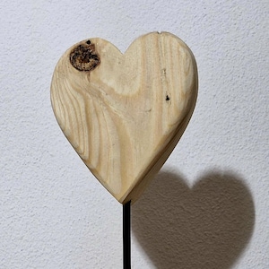 Könnte beinhalten: Holzherz-Skulptur auf einem schwarzen Metallständer. Das Herz besteht aus hellem Holz mit einem Astlochdetail. Der Ständer hat einen runden Sockel und einen schlanken Stiel. Die Skulptur wirft einen herzförmigen Schatten.