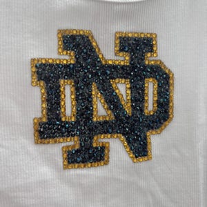 May include: White ribbed vest top with a large, embellished "ND" logo. The logo is outlined in gold and filled with dark blue and teal rhinestones. The vest top is made of a soft, ribbed fabric.