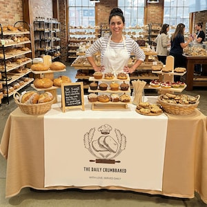 May include: A bakery display featuring a variety of baked goods, including muffins, cookies, and bread. The table is covered with a tan tablecloth and has a sign that says "The Daily Crumbbaked". A woman in a striped shirt and apron stands behind the table.