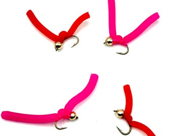 May include: Four fishing flies with curved, worm-like bodies in red and pink. Each fly has a gold-colored bead and a curved hook. The flies are arranged against a white background, showcasing their vibrant colors and design.