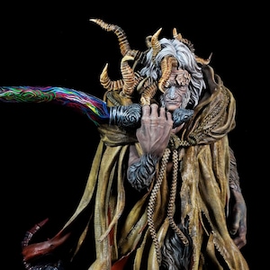 May include: A detailed figurine of a robed figure holding a long, multi-colored blade. The figure has white hair and is adorned with gold accents. The base is a dark gray with ornate carvings. The background is black.