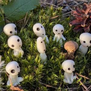 May include: A collection of small, white figurines with black eyes and a mushroom cap, set amidst green moss and a pine cone. The figurines have a smooth, rounded appearance, resembling characters from a fantasy setting.