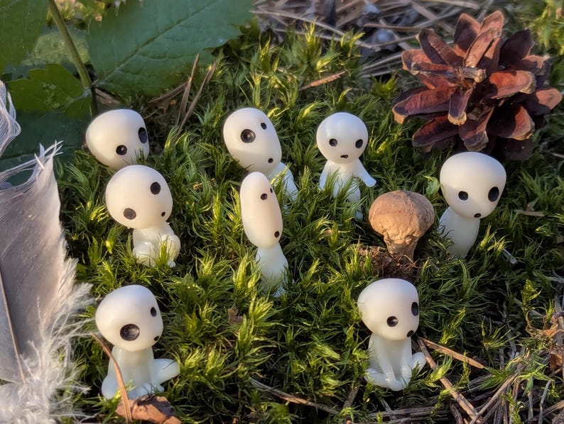 May include: A collection of small, white figurines with black eyes and a mushroom cap, set amidst green moss and a pine cone. The figurines have a smooth, rounded appearance, resembling characters from a fantasy setting.