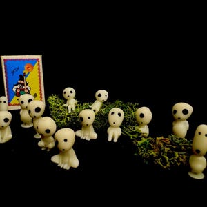 May include: A collection of small, off-white figurines with large, dark eyes, resembling characters from a fantasy film, are arranged on a bed of green moss. A small, colorful card with a cartoon image is in the background.