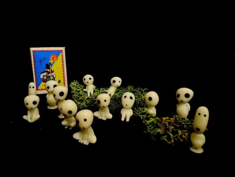 May include: A collection of small, off-white figurines with large, dark eyes, resembling characters from a fantasy film, are arranged on a bed of green moss. A small, colorful card with a cartoon image is in the background.