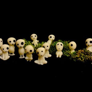 May include: A collection of small, off-white figurines with large, black eyes and simple features. The figures are arranged on a bed of green moss against a black background. Some are standing, others are seated.