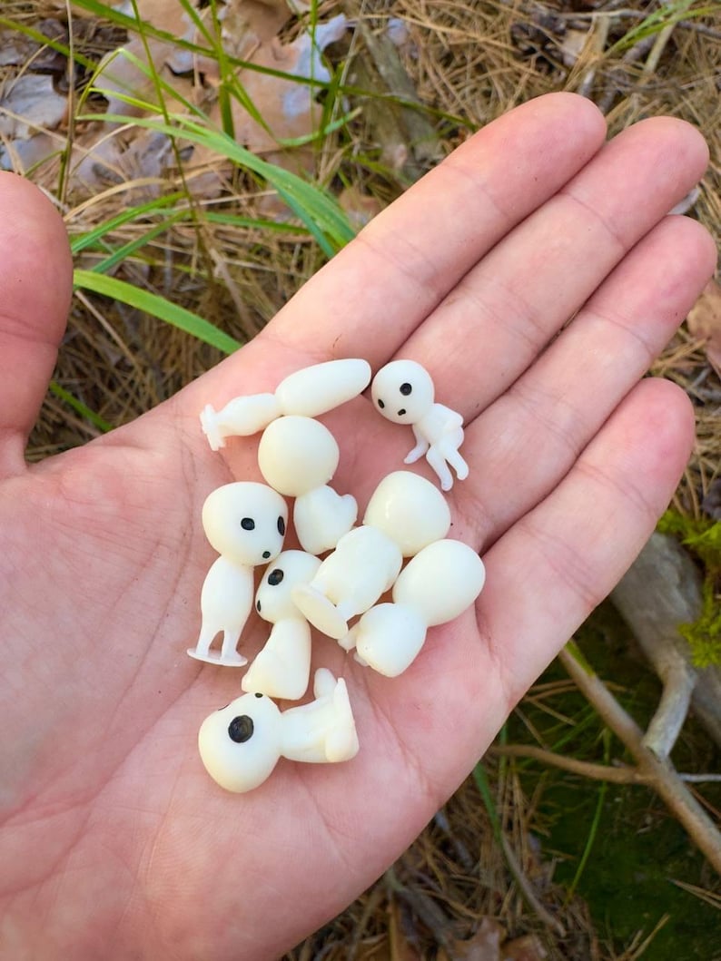 May include: A collection of small, white Kodama figurines with black eyes, held in an open hand. The figurines are in various poses, some standing and others lying down. The background shows dry grass and green foliage.