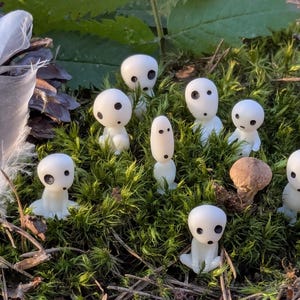 May include: A collection of small, white figurines with black eyes and a mushroom, nestled in green moss. A large white feather and a pine cone are also present. The figurines have a whimsical, fantasy theme.
