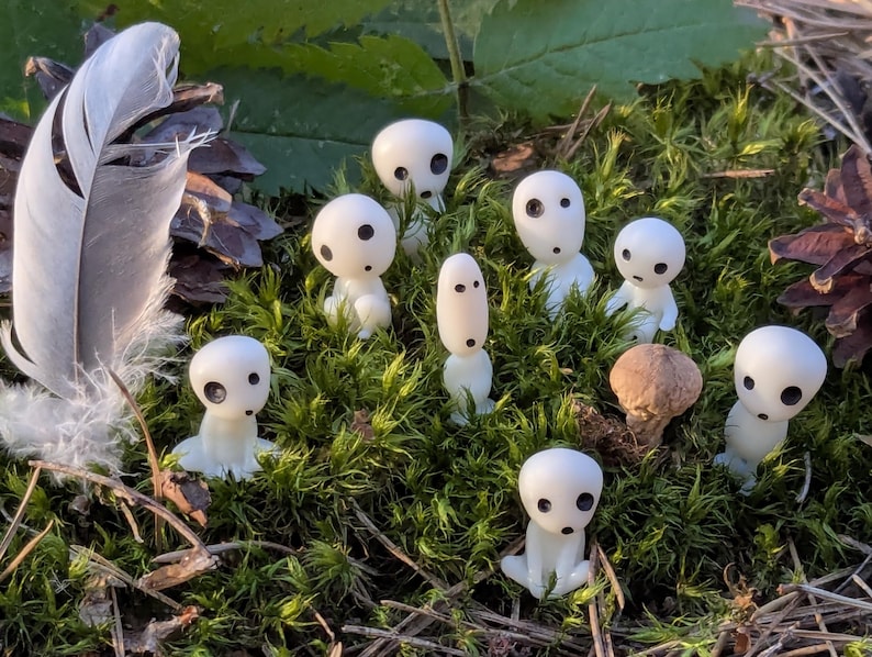 May include: A collection of small, white figurines with black eyes and a mushroom, nestled in green moss. A large white feather and a pine cone are also present. The figurines have a whimsical, fantasy theme.
