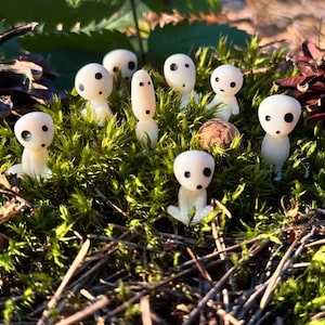 Może przedstawiać: Kolekcja małych, białych figurek z czarnymi oczami i ustami, przypominających leśne duchy, umieszczona wśród zielonego mchu i igieł sosnowych. W tle widoczne są dwa szyszki.