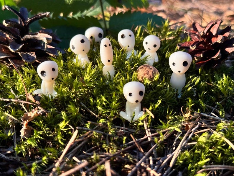 May include: A collection of small, white figurines with black eyes and mouths, resembling forest spirits, are nestled among green moss and pine needles. Two pine cones are visible in the background, creating a natural setting.