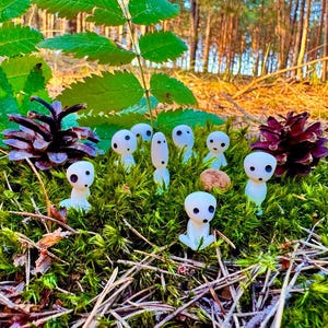 May include: A collection of small, white figurines with black eyes, resembling forest spirits, are nestled in vibrant green moss. The scene includes pine cones and a natural forest backdrop, creating a whimsical, nature-inspired display.
