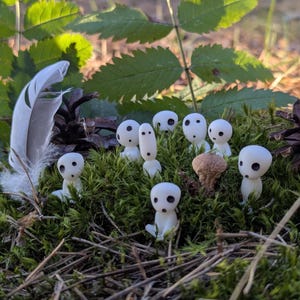 May include: A collection of small, white Kodama figurines with black eye-like markings, nestled in green moss. A feather and pine cone are also present, creating a natural, forest-like scene.