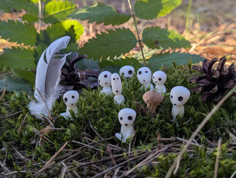 May include: A collection of small, white Kodama figurines with black eye-like markings, nestled in green moss. A feather and pine cone are also present, creating a natural, forest-like scene.