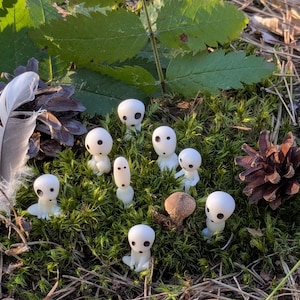May include: A collection of small, white figurines with black eyes, resembling forest spirits, are scattered on a bed of green moss. A feather, pine cone, and a small mushroom are also present, creating a natural woodland scene.