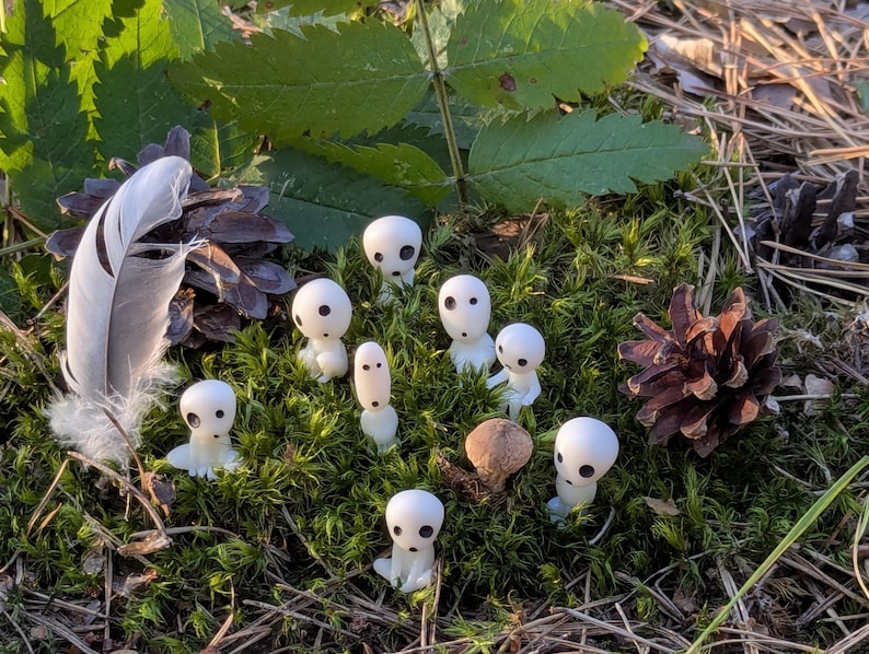 May include: A collection of small, white figurines with black eyes, resembling forest spirits, are scattered on a bed of green moss. A feather, pine cone, and a small mushroom are also present, creating a natural woodland scene.