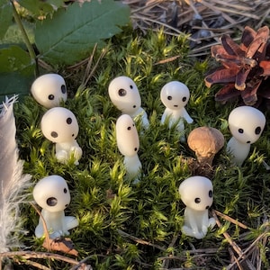 May include: A collection of small, white figurines with black dot eyes, resembling forest spirits, are arranged on a bed of green moss. A feather and pine cone are also present, creating a natural, whimsical scene.