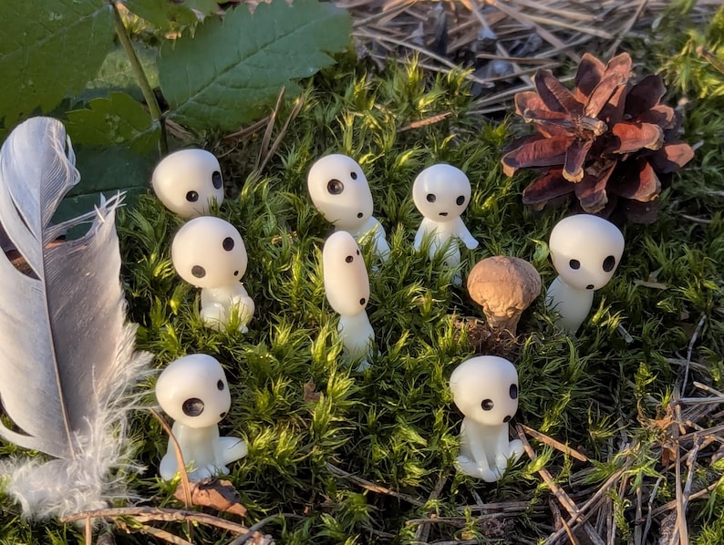 May include: A collection of small, white figurines with black dot eyes, resembling forest spirits, are arranged on a bed of green moss. A feather and pine cone are also present, creating a natural, whimsical scene.
