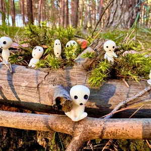 May include: A collection of small, white figurines with black eyes and mouths, resembling forest spirits, are arranged on a weathered log with green moss. The figurines are set against a backdrop of a sunlit forest.