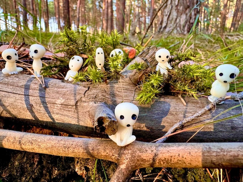 May include: A collection of small, white figurines with black eyes and mouths, resembling forest spirits, are arranged on a weathered log with green moss. The figurines are set against a backdrop of a sunlit forest.