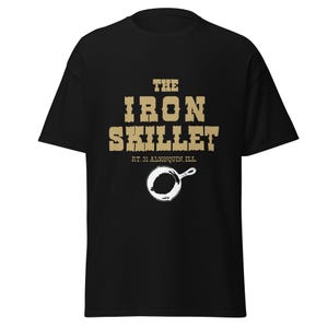 May include: Black t-shirt with a gold-colored graphic that reads "THE IRON SKILLET" above "RT. 31 ALGONQUIN, ILL" and a white skillet illustration. The shirt is made of a soft, comfortable material.