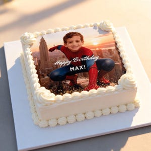 May include: Square white cake with a Spiderman image and the text "Happy Birthday MAX!". The cake is decorated with white frosting and a border of piped rosettes. The Spiderman image features a boy in a Spiderman costume.