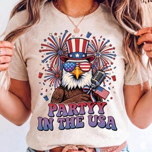 May include: A beige t-shirt featuring a graphic of a bald eagle wearing a patriotic top hat and sunglasses. The eagle is next to a microphone and a small American flag. Fireworks, red cups, and the text "PARTYY IN THE USA" are printed below.