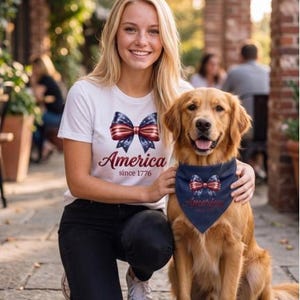 May include: A white t-shirt featuring a red, white, and blue bow graphic with the text America since 1776, paired with a matching navy blue dog bandana that displays the same bow graphic and the text America since 1776 in red lettering.