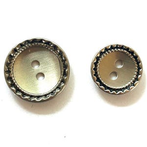 May include: Two round metallic buttons with a brushed gold-tone centre and a decorative scalloped edge. Each button features two central holes for attachment. The buttons are of different sizes, with one being noticeably larger than the other.