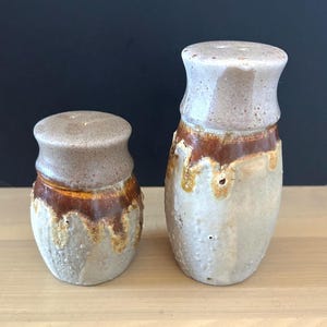 May include: Two ceramic salt and pepper shakers with a textured, light gray base and a smooth, tan-colored top section. A dark brown glaze drips from the top rim down onto the textured body, creating a distinct contrast between the two finishes.