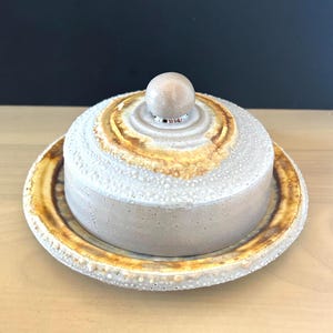 May include: A round ceramic butter dish with a lid featuring a central spherical knob. The piece has a textured, speckled light gray finish with prominent rings of warm amber and brown glaze around the lid and the outer edge of the base plate.