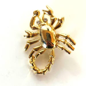 May include: A gold-toned scorpion brooch featuring a polished, reflective body and two small, clear, circular rhinestones set as eyes. The scorpion has articulated legs and a curved tail, with a smooth, shiny finish across the entire metal surface.