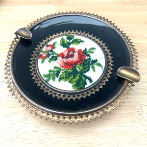May include: A round ashtray featuring a black glass center with a cross-stitch floral design of a red rose and green leaves. The ashtray is framed by a decorative gold-tone metal border with a scalloped edge and two integrated metal rests.