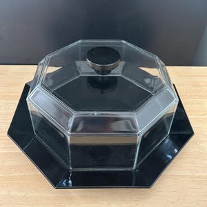 May include: A black and clear glass serving dish with a lid. The dish has an octagonal shape with a black base and a clear glass lid with a black knob. The dish is sitting on a wooden surface.