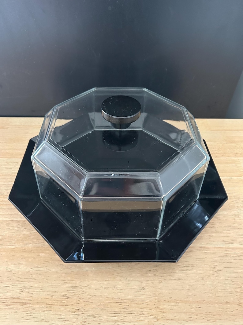 May include: A black and clear glass serving dish with a lid. The dish has an octagonal shape with a black base and a clear glass lid with a black knob. The dish is sitting on a wooden surface.