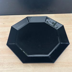 May include: A black, octagonal plate with a glossy finish. The plate is set on a light-colored wooden surface. The plate's geometric design adds a modern touch to any table setting.
