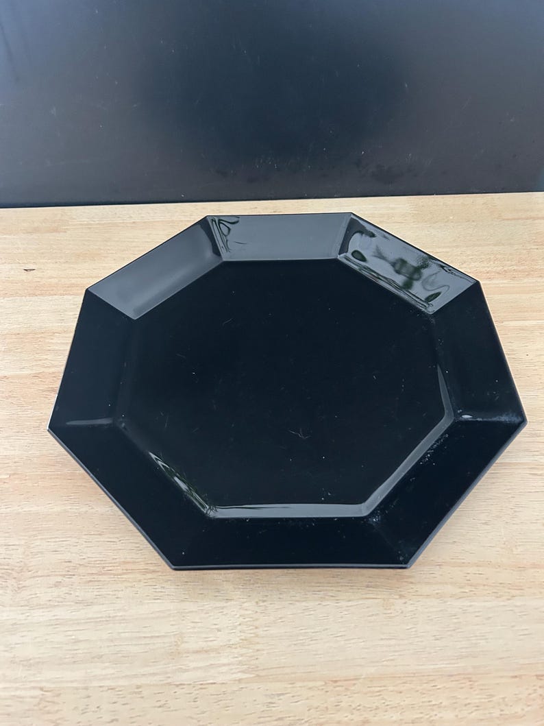May include: A black, octagonal plate with a glossy finish. The plate is set on a light-colored wooden surface. The plate's geometric design adds a modern touch to any table setting.