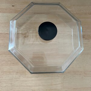 May include: Clear, octagonal glass container with a black, circular lid. The container is empty and sits on a light brown wooden surface. The lid is centered inside the container.