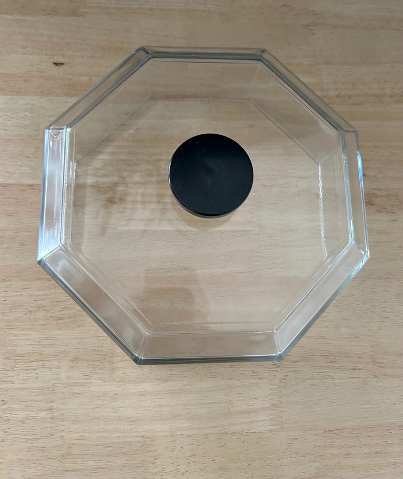 May include: Clear, octagonal glass container with a black, circular lid. The container is empty and sits on a light brown wooden surface. The lid is centered inside the container.