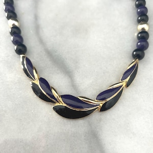 May include: A vintage necklace featuring a gold-tone leaf design with black and navy blue enamel accents. The necklace has a beaded chain with black, navy blue, and gold-tone beads. A stylish accessory for any outfit.