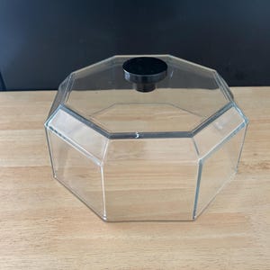 May include: A clear, octagonal container with a black knob on the lid. The container is made of transparent material, allowing visibility of the contents. The lid is a solid black color. The container sits on a light-colored wooden surface.