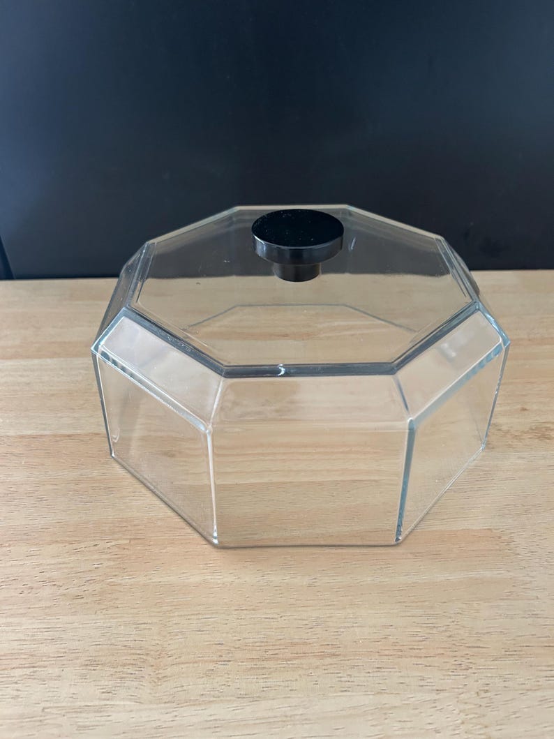 May include: A clear, octagonal container with a black knob on the lid. The container is made of transparent material, allowing visibility of the contents. The lid is a solid black color. The container sits on a light-colored wooden surface.