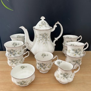 May include: A white porcelain tea set with a floral design. The set includes a teapot, creamer, sugar bowl, and several teacups. The floral design features grey and yellow roses with gold trim. The teacups are stacked in groups.