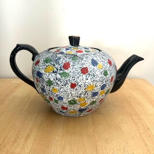 Puede incluir: Una tetera de cerámica con un cuerpo moteado gris y blanco, adornada con lunares de colores rojo, amarillo, azul y verde. Cuenta con un asa, un pico y una tapa negros con un pequeño pomo negro. La tetera está sobre una superficie de madera.
