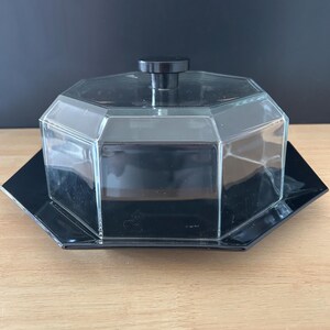 May include: A clear, octagonal glass butter dish with a black lid and base. The lid has a small, round black handle. The dish sits on a black, octagonal base, creating a sleek, modern look. The butter dish is on a wooden surface.