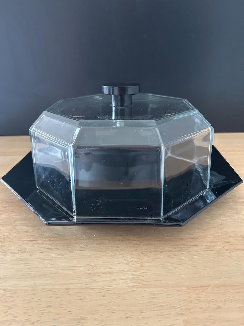 May include: A clear, octagonal glass butter dish with a black lid and base. The lid has a small, round black handle. The dish sits on a black, octagonal base, creating a sleek, modern look. The butter dish is on a wooden surface.